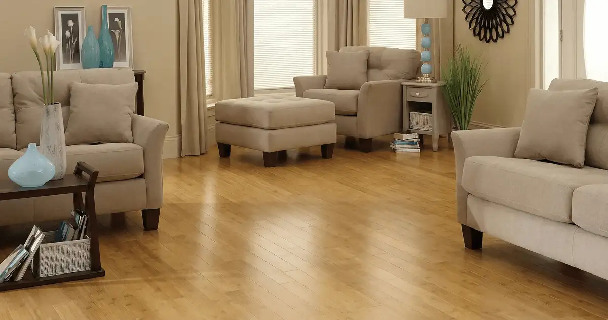 A living room with professionally installed bamboo flooring, beige sofas, a matching ottoman, a side table with a blue lamp, vases, framed pictures, and large windows with blinds and curtains in Orlando, FL